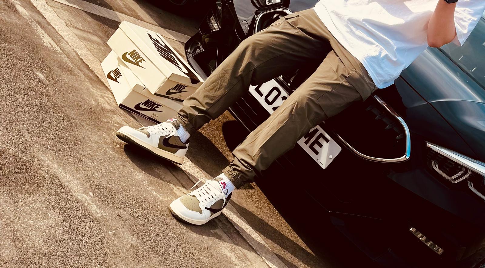 Close-up of replica Travis Scott Jordan 1 Low Reverse Olive trainers worn by a model leaning against a car—highlighting premium sneaker dupes with signature backward Swoosh, earthy tones, and affordable designer-inspired streetwear style.